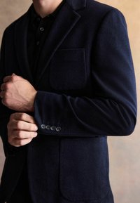 Marinblå ullblazer med en enda bröstficka, sexknappsdetalj vid ärmslutet och textilstruktur. Modellen justerar ärmslutet.