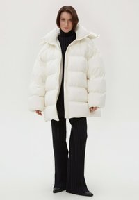 Biała puchowa kurtka z wysokim kołnierzem i oversize'owym krojem, z pikowanymi panelami. Modelka ma na sobie ciemne, prążkowane spodnie i zaostrzone buty.