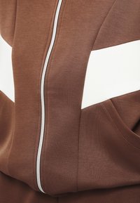 Gros plan d'une veste marron à fermeture éclair avec une bande horizontale blanche et une poche latérale, montrant la texture du tissu et les détails des coutures.