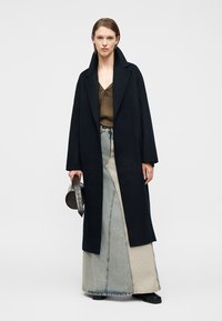 Sieviete valkā garu melnu mēteli, olīvu blūzi, divu toņu džinsu maxi svārkus, melnus apavus un tur apaļu melnu somu.