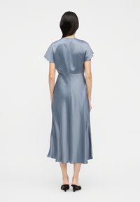 Γυναίκα με μακριά σκούρα  μαλλιά, ντυμένη με ανοιχτό γαλάζιο σατέν μίντι φόρεμα και μαύρα ψηλοτάκουνα mules, που στέκεται με την πλάτη στραμμένη σε ένα απλό φόντο.