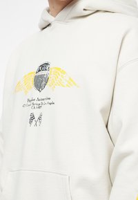 Krémová mikina s kapucí s grafickým potiskem na přední straně, který zobrazuje design štítu, žlutá křídla a text. Obsahuje klokaní kapsu a jemnou texturu.