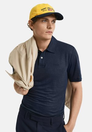 Polo shirt blu navy con tessuto testurizzato, abbinato a un giubbotto beige lasciato cadere sulla spalla. Il modello indossa un cappello giallo con dettagli del logo.