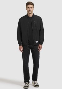 Schwarze Bomberjacke mit seitlichen Taschen und einer Reißverschlusstasche auf der Brust, getragen über einem schwarzen Hemd, kombiniert mit dunklen Jeans und schwarz-weißen Turnschuhen.