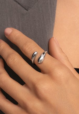 Conjunto de anillos de plata de doble banda con formas lisas y curvas, con un acabado pulido y diseño ligeramente superpuesto, sobre una mano apoyada en una tela.