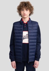 Gilet matelassé bleu marine avec texture piquée, fermeture éclair à l'avant, col haut, porté sur un sweat bleu marine avec des accents rouges et un logo.