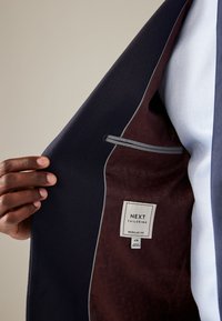 Tumesinine bleiser tekstuurse kangaga, millel on burgundia toonis vooder, halli ääristus ja silt, millel on kiri "NEXT TAILORING REGULAR FIT".