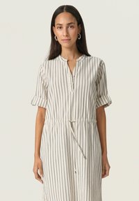 Robe légère beige avec des rayures verticales bleu marine, manches courtes retroussées, encolure ronde et un détail de taille à cordon.