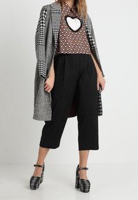 Cappotto a scacchi grigio e nero sopra un top marrone a fantasia con un taglio a cuore, abbinato a pantaloni cropped neri e tacchi a zeppa a scacchi neri.