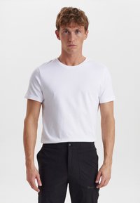 Hvid kortærmet t-shirt med rund halsudskæring, lavet af et glat stof, båret med sorte bukser med sidelommer og en knap.