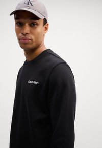 Felpa nera con scollo rotondo, con il logo bianco "Calvin Klein" sul petto. Il modello indossa un cappellino grigio chiaro. Tessuto morbido.