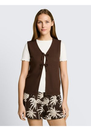 Junge Frau trägt ein weißes kurzärmeliges Hemd, eine braune ärmellose Weste mit Schleifen und braune Shorts mit weißem Palmenmuster.