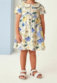 Niña vestida con un vestido crema con patrones florales azules y amarillos, y un parche de flor morada sonriente, combinado con sandalias blancas.