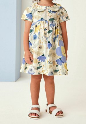 Enfant portant une robe crème avec des motifs floraux bleus et jaunes et un patch de fleur souriante violet, associé à des sandales blanches.