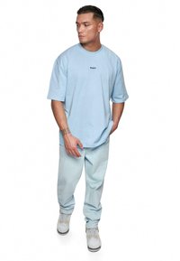 Lyseblå oversized T-shirt med et lille sort logo ved brystet, parret med lyse denimbukser og hvide sneakers.
