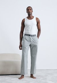 Balta bezmaku kreklu un pelēkajām svītrainajām pidžamu biksēm ar melnu jostu, uz kuras ir "BJÖRN BORG" logo, valkā stāvošs vīrietis gaišā paklājā.