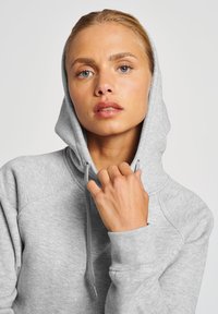 Szara bluza z kapturem, wykonana z miękkiego materiału, z kapturem na sznurku, rękawami raglanowymi i ściągaczami przy mankietach. Model trzyma kaptur blisko twarzy.