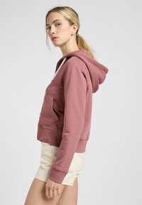 Sudadera rosa hecha de una tela suave, con un bolsillo tipo canguro, puños acanalados y capucha con cordón. Combinada con pantalones cortos de color claro.