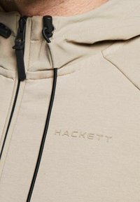 Sudadera con capucha beige de textura suave, que presenta herrajes de cremallera negra y un logo sutil de "HACKETT" estampado en la tela.
