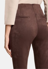 Bruine suède legging met een gladde textuur, een hoge tailleband en twee achterzakken. Close-up weergave van de stof en pasvorm.