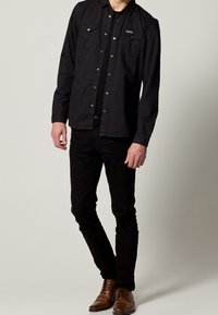 Camicia nera con bottoni e due tasche sul petto, abbinata a jeans neri slim-fit e scarpe eleganti marroni. Design e texture minimalisti.