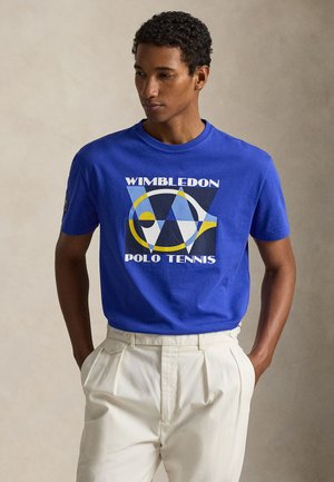 Άνδρας που φορά μπλε μπλουζάκι "Wimbledon Polo Tennis" και λευκό παντελόνι, στέκεται με τα χέρια στις τσέπες, μπροστά σε ουδέτερο φόντο.