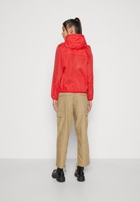 Chaqueta ligera roja con capucha, puños elásticos, combinada con pantalones cargo beige que tienen bolsillos laterales y un corte recto. Botas negras gruesas.