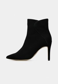 Botín negro con punta afilada y un diseño curvado. Hecho de material suave, tiene un alto tacón de aguja y una suela beige.