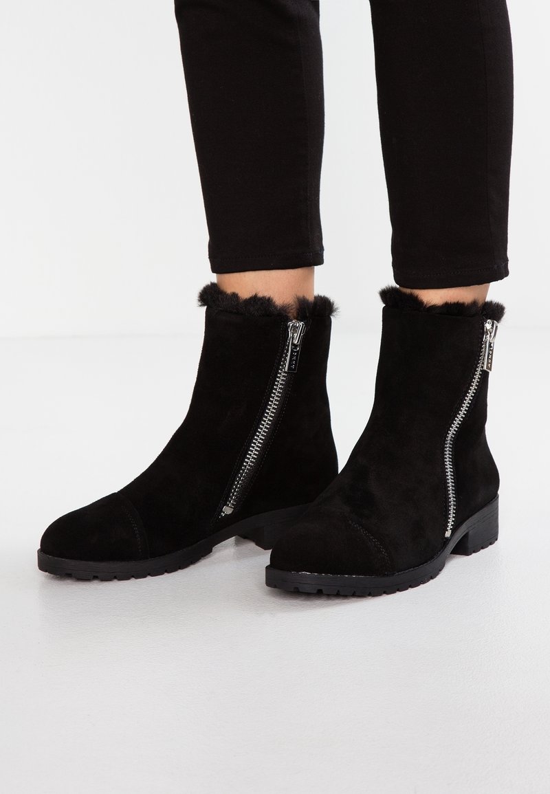 Les bottines en daim noir présentent une fermeture éclair de chaque côté, une bordure en fausse fourrure en haut, et une semelle en caoutchouc plate avec une prise texturée.