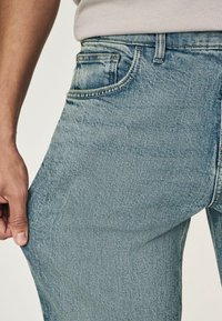 Jeans en denim bleu clair avec une texture lisse, un design à cinq poches et une couture visible. Le tissu semble légèrement usé par endroits.
