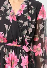 Robe portefeuille en tissu noir ornée de grandes fleurs roses, de feuilles vertes et d'un détail ceinturé à la taille. Manches longues transparentes.
