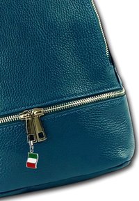 Teal leren handtas met een gouden rits en een klein Italiaans vlaggetje eraan bevestigd. Heeft een gestructureerd oppervlak en een afgeronde bodem.