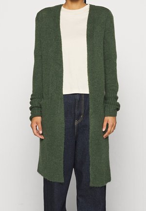Personne portant un long cardigan tricoté vert foncé ouvert sur le devant, par-dessus une chemise blanche unie et un jean bleu foncé, debout devant un fond uni.