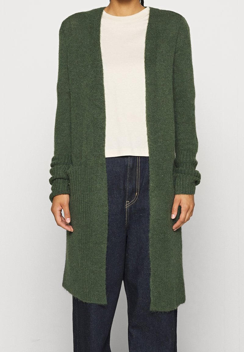 Personne portant un long cardigan tricoté vert foncé ouvert sur le devant, par-dessus une chemise blanche unie et un jean bleu foncé, debout devant un fond uni.