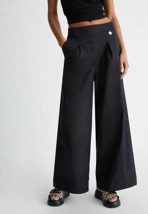 Osoba nosząca czarne szerokie spodnie z plisami i przypinką w kształcie gwiazdy, w połączeniu z sandałami na platformie w panterkę, stojąca z jedną ręką w kieszeni.