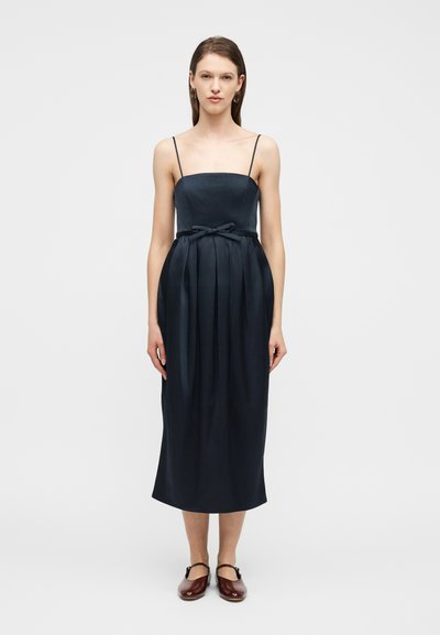 Femme portant une robe midi bleu marine à fines bretelles, un petit nœud à la taille, et des chaussures Mary Jane bordeaux, debout sur un fond blanc.