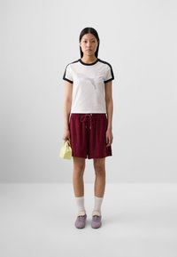A young woman stands against a minimalistic background, wearing a white T-shirt with a Puma logo, maroon shorts, and purple sneakers, holding a small yellow bag.