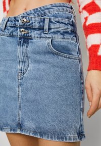 Mini-jupe en denim bleu clair, dotée d'une taille haute avec deux fermetures par boutons, de poches avant et de détails subtils d'usure.