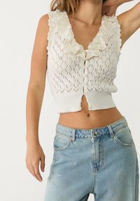 Biały, dziany top w stylu crop z diamentowym wzorem, falbaniastym dekoltem i zapięciem na guziki z przodu. W połączeniu z jasnoniebieskimi spodniami z wysokim stanem.