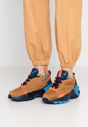 Persona con pantalones caqui remangados y zapatillas Reebok marrones, azul marino, naranja y azul, de pie sobre una superficie lisa y clara.