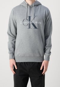 Hombre vistiendo una sudadera gris de Calvin Klein con bolsillo frontal y pantalones negros, de pie contra un fondo claro y liso.