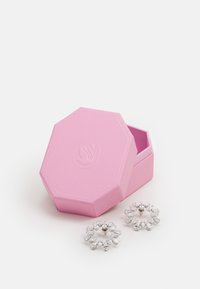 Caja de regalo octagonal rosa con exterior texturizado, ligeramente abierta, revelando dos aretes circulares adornados con diversas piedras transparentes.