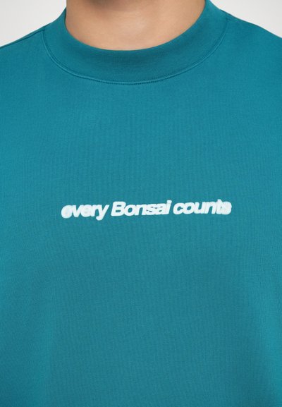 Tirkizna pamučna majica s okruglim izrezom; sadrži bijeli, blago uzdignuti tekst "svaki Bonsai se računa" centriran na prsima.