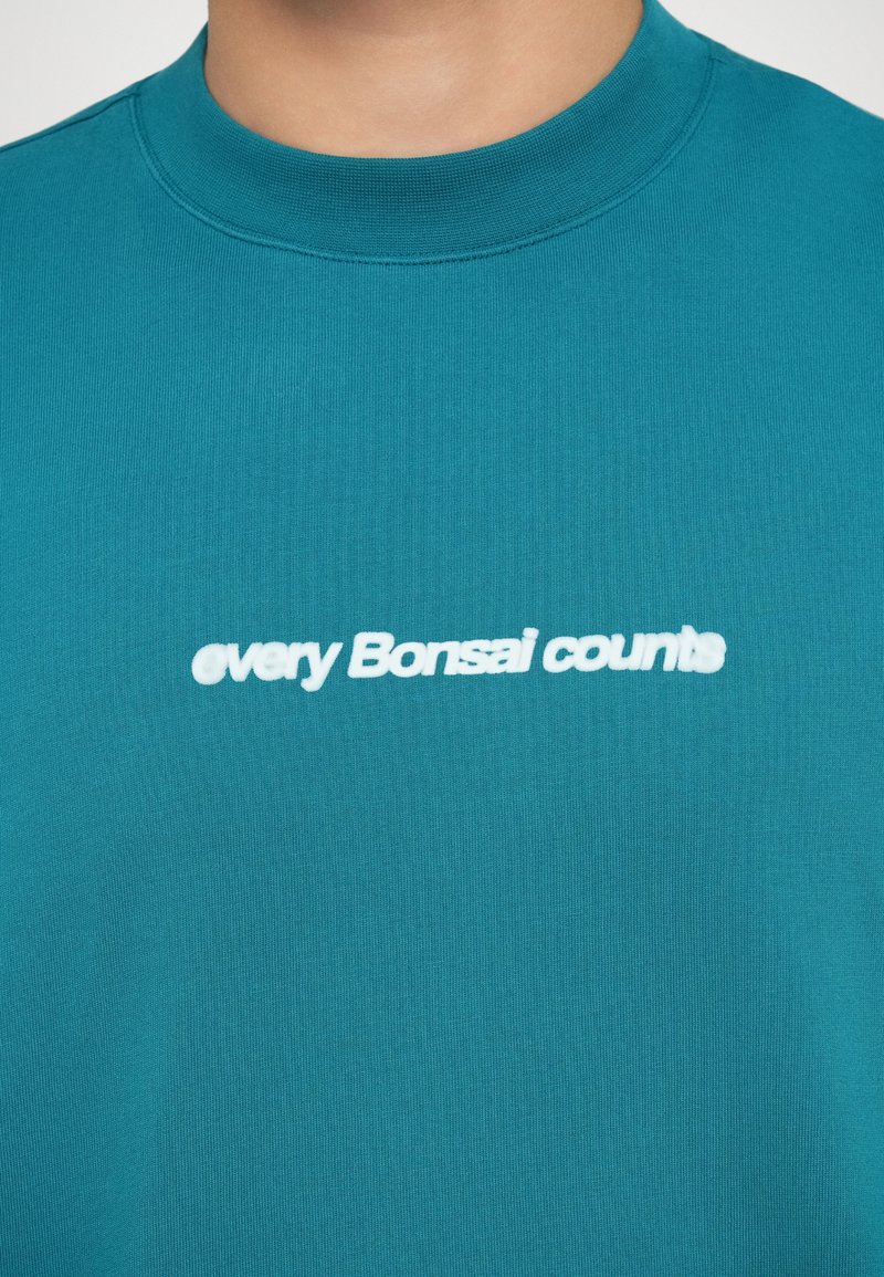 T-shirt en coton teal avec un col rond ; présente un texte blanc, légèrement en relief, indiquant "chaque Bonsai compte" centré sur la poitrine.