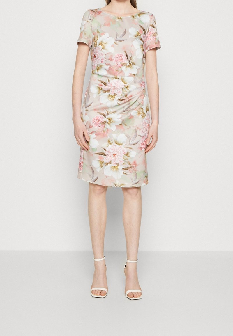 Femme portant une robe courte à manches courtes avec un motif floral rose et blanc et des sandales blanches à talons hauts avec bride à la cheville.