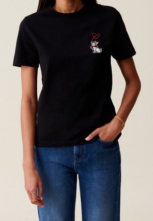 Kobieta w czarnej koszulce z nadrukiem małego białego psa i czerwonego serca na piersi, w niebieskich dżinsach i złotych bransoletkach.