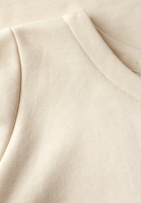 Béžová bavlněná tkanina zblízka, s žebrovanou texturou a kulatým výstřihem. Měkká, lehká s hladkým povrchem a bez viditelných vzorů.