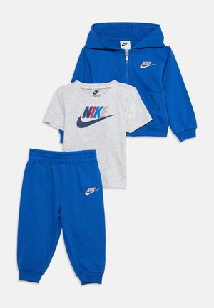 Niebieska bluza z kapturem i zamkiem, szary t-shirt Nike z krótkim rękawem i wielokolorowym logo oraz niebieskie spodnie dresowe Nike dla dzieci, ułożone płasko na białym tle.