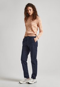 Strikket beige genser med høy hals og diamantmønster, parret med marineblå bukser. Modellen har på seg grå joggesko.