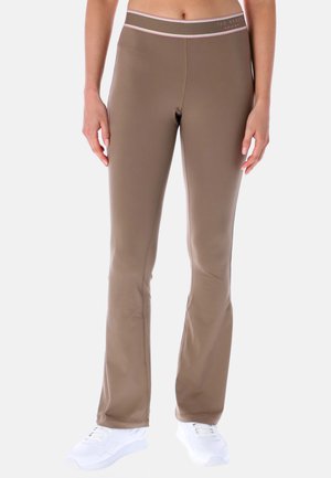 Femme portant un pantalon évasé marron cintré et des baskets blanches, debout, les bras détendus le long du corps, sur un fond clair uni.